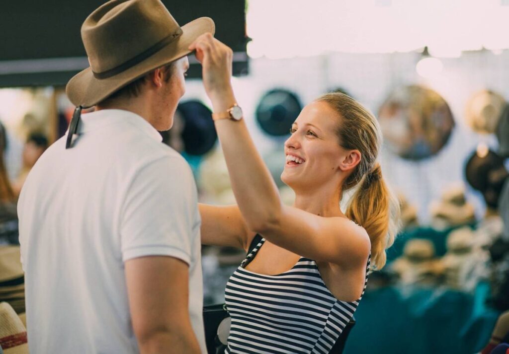 Young couple are exploring Queen Victoria Market in Australia. The woman is putting a hat on her boyfriend in a market stall. (Young couple are exploring Queen Victoria Market in Australia. The woman is putting a hat on her boyfriend in a market stall